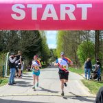 DSC_6255 Biegacze startują spod różowego łuku startowego. Tło to zielone drzewa i jasne niebo. Naokoło stoją kibice.