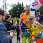 DSC_6264 Na zdjęciu para biegaczy w kolorowych strojach z medalami. W tle widoczne są drzewa i baner startowy. Pogoda jest słoneczna, co dodaje energii wydarzeniu.