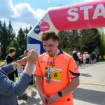 DSC_6274 Biegacz w pomarańczowej koszulce otrzymuje medal. Tło to zielone drzewa i niebo z chmurami. Nad nim widoczny jest łuk startowy.