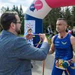 DSC_6290 Na zdjęciu mężczyzna w niebieskim stroju biegowym otrzymuje medal. W tle znajduje się brama z napisem Ale czas! i drzewa. Obok widoczna jest grupa kibiców.