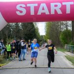 DSC_6297 Dwie kobiety biegną pod różowym łukiem startowym na asfaltowej drodze. W tle zielone drzewa i widzowie. Kobieta w czarnej koszulce numer 1, druga w niebieskiej numer 24.