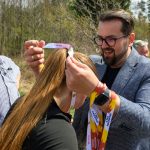DSC_6313 Na zdjęciu mężczyzna zakłada medal na szyję młodej kobiety. Tło stanowi las, a atmosfera jest słoneczna. Medale mają jasne kolory z napisami.