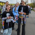 DSC_6317 Grupa młodych osób trzyma medale, stojąc na asfaltowej drodze. Tło to zielone drzewa i niebo z chmurami. Dominują ciemne i jasne ubrania.