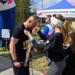 DSC_6319 Biegacz otrzymuje medal od kobiety przy mecie. Tło zielone z niebieskim namiotem i zegarem z czasem 0:42:16.