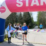 DSC_6322 Biegaczka przekracza linię mety pod różowo-białym łukiem startowym. Na poboczu stoją ludzie i kibicują. W tle drzewa i asfaltowa droga.