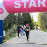 DSC_6334 Biegaczka w czarnym stroju przekracza linię startu pod różowym banerem „START”. W tle drzewa i kibice. Pogoda jest słoneczna.