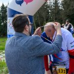 DSC_6344 Mężczyzna zakłada medal biegaczowi na mecie. W tle widać las i kilku obserwatorów. Ubrania w głównie niebiesko-białych kolorach.