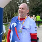 DSC_6347 Mężczyzna w sportowym stroju z medalem stoi na zawodach biegowych. Ubrany w biało-niebieską koszulkę z czerwonymi akcentami. W tle widoczne są drzewa i ludzi.
