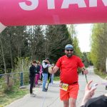 DSC_6348 Zawodnik w czerwonym stroju kończy marsz nordic walking pod różowym balonem START na leśnej drodze. W tle inni uczestnicy i drzewa. Pogoda jest słoneczna.