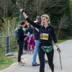 DSC_6352 Na zdjęciu kobieta z numerem startowym 186 kończy nordic walking. Tło to drzewa i grupa ludzi. Przewaga kolorów zielonego i czarnego.