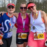 DSC_6356 Trzy uśmiechnięte osoby w sportowych strojach ze srebrnymi medalami. Kolory: różowy, biały, niebieski. W tle drzewa i uczestnicy biegu.