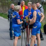 DSC_6359 Na zdjęciu grupa osób w sportowych strojach w niebieskim kolorze stoi na drodze. Trzymają kijki do nordic walking, a w tle widać drzewa. Atmosfera jest luźna, a w tle są inni uczestnicy.