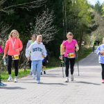 DSC_6363 Grupa ludzi biegnie po brukowanej ścieżce przez las. Kobieta w różowej koszulce używa kijków do nordic walking. Tło to zieleń drzew i jasne niebo.