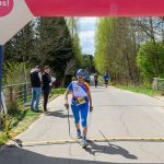 DSC_6373 Zawodniczka na trasie biegu w niebieskiej odzieży sportowej. W tle wiosenna zieleń drzew. Napis Ale czas! na łuku nad drogą.