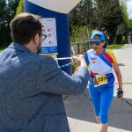 DSC_6376 Starsza kobieta w niebieskim stroju biegnie na zawodach. Mężczyzna w marynarce wręcza jej kartkę. W tle las i droga.
