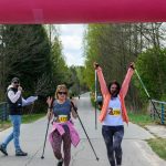 DSC_6394 Dwie kobiety z kijkami przekraczają linię startu na zawodach nordic walking. Tło to zielony las i droga. Nad nimi różowy napis START.