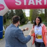DSC_6399 Na zdjęciu kobieta z numerem startowym 198 kończy bieg w parku. Jest uśmiechnięta i trzyma kijki do nordic walking. W tle znajduje się łuk startowy i zalesiona ścieżka.