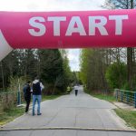 DSC_6401 Zdjęcie przedstawia start biegu pod różowo-białym łukiem z napisem START. W tle widoczny jest las i kilka osób na drodze. Po prawej stronie znajduje się wyświetlacz z czasem.