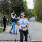 DSC_6403 Na zdjęciu widoczna jest kobieta w białej koszulce i okularach, korzystająca z kijków do nordic walking. W tle znajdują się drzewa i dwie osoby z mikrofonem i kartkami. Nad nią rozciąga się różowy łuk, na drodze asfaltowej.