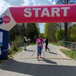 DSC_6410 Na zdjęciu osoba w kolorowym stroju przechodzi przez linię startu biegu w lesie. Nad nią dmuchany łuk w kolorach różowym i niebieskim z napisem START. Obok widoczny jest zegar z czasem, a w tle drzewa i droga.