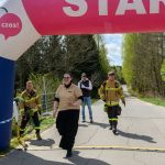 DSC_6413 Na zdjęciu jest start biegu w plenerze z nadmuchiwanym łukiem w kolorach różowym i niebieskim. Strażacy idący po asfalcie, otoczeni są zielenią drzew. Obok łuku widoczna jest mapa Polski z zaznaczonymi miejscami.