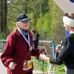 DSC_6424 Na zdjęciu dwie osoby rozmawiają na zewnątrz. Osoba po lewej ma czerwony polar, czapkę i medal. Tło to zielone drzewa i niebieska konstrukcja.