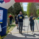 DSC_6428 Grupa ludzi z numerami startowymi kończy marsz nordic walking. Pod nadmuchanym łukiem widać zieleń i słoneczne niebo. W tle las i wąska droga.
