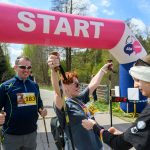 DSC_6431 Start wyścigu nordic walking z różowym łukiem na tle zieleni. Kobieta z kijami unosi ręce w geście triumfu. Obok niej mężczyzna i wolontariuszka.