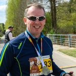 DSC_6434 Na zdjęciu mężczyzna w okularach przeciwsłonecznych na tle zielonych drzew. Nosi niebieską kurtkę sportową i medal. W tle widać metalowe ogrodzenie i drogę.
