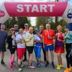 DSC_6458 Grupa biegaczy z medalami stoi pod różowo-białym łukiem z napisem START. W tle drzewa i niebo. Uczestnicy są uśmiechnięci.