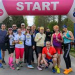 DSC_6465 Grupa osób w kolorowych strojach sportowych stoi pod różowym łukiem START. Tło z zielonymi drzewami. Wszyscy trzymają medale.
