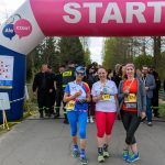 DSC_6470 Trzy kobiety z medalami stoją przy różowej bramie startowej. Tło z zielonymi drzewami i ludźmi w strojach sportowych. Obok znajduje się zegar z czasem.