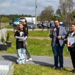 DSC_6488 Na zdjęciu jest kilka osób na świeżym powietrzu. Mężczyzna przemawia do mikrofonu, a kobieta trzyma dyplom. Tło to zielona trawa, drzewa i samochody.