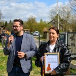 DSC_6490 Na zdjęciu jest mężczyzna z mikrofonem i kobieta z ramką. Tło to zielona trawa i niebo z chmurami. Kobieta trzyma dyplom z podziękowaniem.