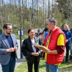 DSC_6498 Na zdjęciu widać grupę ludzi na zewnątrz, przy mikrofonie. Tło stanowią drzewa i ogrodzenie, kolory są jasne i wiosenne. Osoby są uśmiechnięte, a atmosfera jest pozytywna.