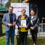 DSC_6501 Trzy osoby stoją na trawie przed banerem z napisem Mazowsze. Jedna z nich trzyma certyfikat. Niebo jest błękitne i bezchmurne.