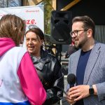 DSC_6504 Trzy osoby rozmawiają na zewnątrz. Kobieta w różowej bluzie z nadrukiem Ale czas!. Mężczyzna trzyma mikrofon; tło jest zielone.
