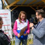 DSC_6507 Na obrazie są trzy osoby na zewnątrz. Kobieta w biało-różowej bluzie trzyma certyfikat, rozmawiając z mężczyzną z mikrofonem. W tle jest baner Mazowsze i drewniana konstrukcja.