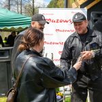 DSC_6516 Na zdjęciu troje ludzi, w tym dwóch w mundurach, stoi na trawie. W tle zielony namiot i biały baner Mazowsze. Osoba wręcza coś drugiej.