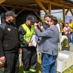 DSC_6520 Na zdjęciu widać ceremonię wręczenia nagród na zewnątrz. Osoba w kamizelce odbiera medal, stojąc przed innymi ludźmi. W tle widoczne są tła z logotypem Mazowsze i podium.