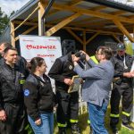 DSC_6523 Grupa strażaków otrzymuje nagrody na tle drewnianej altanki. W prawym górnym rogu widać baner Mazowsze. Strażacy ubrani są w czarne uniformy z żółtymi elementami.