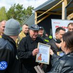 DSC_6528 Grupa osób w mundurach otrzymuje dyplom, tło zielone drzewo i namiot. Mężczyzna trzyma ramkę, obok mikrofon. Pogodne niebo.