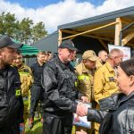 DSC_6535 Grupa strażaków w czarnych i żółtych mundurach stoi na zewnątrz, przy namiocie. Jeden z nich podaje dokument kobiecie w skórzanej kurtce. Tło stanowi budynek z drewna i zielony namiot.