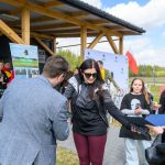 DSC_6554 Grupa ludzi na zewnątrz, kobieta otrzymuje nagrodę. Drewniana konstrukcja w tle, widoczna polska flaga. Słoneczny dzień, niebo z białymi chmurami.