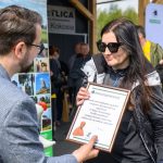 DSC_6556 Na zdjęciu kobieta w okularach przeciwsłonecznych trzyma dyplom podziękowania. Mężczyzna wręcza jej nagrodę. Tło jest rozmyte, z ludźmi oraz banerami.