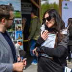 DSC_6558 Na zdjęciu dwoje ludzi rozmawia na zewnątrz. Kobieta w czarnej kurtce trzyma tablicę z tekstem. Tło jest zielone, z plakatami i ludźmi.