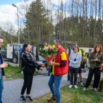 DSC_6582 Grupa ludzi na zewnątrz, mężczyzna wręcza kobiecie bukiet kwiatów. Tło to drzewa i ogrodzenie. Ubrania w żywych kolorach.