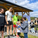DSC_6585 Na zdjęciu trzech mężczyzn na podium otrzymujących nagrody. Ubrani w sportową odzież, w tle drewniana altana i niebo z chmurami. Obok stoją dzieci z pucharami.