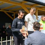 DSC_6587 Trzej mężczyźni z trofeami na podium. Jeden jest w czarnej bluzie, drugi w białej koszulce, trzeci w zielonej kurtce. W tle drewniane zadaszenie i osoby w ubraniach z napisem STRAŻ.