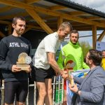 DSC_6588 Trzech mężczyzn na podium, trzymają trofea. Jeden w jasnej koszulce ściska rękę organizatora. Tło z drewnianym zadaszeniem i flagą Polski.